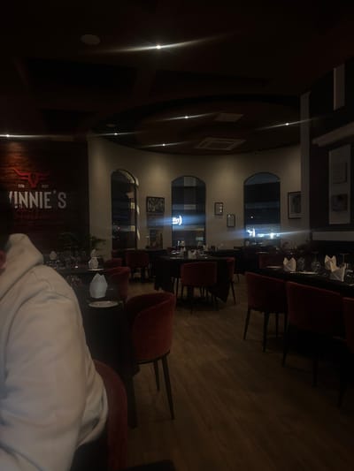 The image displays the dimly lit interior of Vinnie's Steakhouse, located in Spanish Town, British Virgin Islands. The establishment features a dark wood-paneled left wall with a large, illuminated "ESTD 2020 VINNIE'S Steakhouse" logo. The flooring consists of light-toned wood planks.
In the immediate foreground, to the left, the back and upper left shoulder of a person wearing a light-colored hooded garment are visible, seated at a table.
The main dining area is furnished with multiple tables draped in dark (likely black) tablecloths. Each table is set with white folded napkins, wine glasses, and water glasses. Accompanying these tables are numerous chairs upholstered in a reddish-orange or burgundy velvet fabric with dark legs.
The rear wall is a light-colored, curved surface punctuated by several arched windows, through which a nocturnal urban scene is visible, characterized by streetlights and reflections. Framed artwork or decorative items are mounted on this wall between the windows, and also on the dark-paneled wall to the right. A large potted plant is situated near a table on the left side of the room. The dark ceiling contains several recessed spotlights, which produce visible light flares, and an air conditioning vent. No active interactions or significant movements are discernible from any individuals beyond the partially visible person. The restaurant appears to be largely unoccupied.