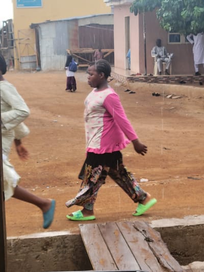 The image captures a scene in Madalla, Nigeria. A woman in a pink and white polka dot top, floral pants, and green sandals walks on a dirt path. Another person wearing a white outfit is partially visible at the bottom. Buildings and other people are visible in the background.