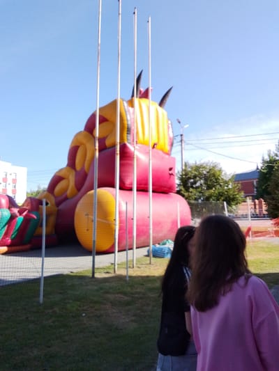 A bright, sunny day in Zavodoukovsk, Russia, showcases an inflatable playground structure, possibly a dragon or monster, colored predominantly in pink and yellow. The structure dominates the background, with a blue sky overhead. In the foreground, two young females are visible, their backs turned to the camera. One wears a pink sweatshirt, while the other is dressed in a black t-shirt and a denim skirt. They are standing on a grassy area adjacent to the playground, separated from it by a fence. The scene appears to be an outdoor recreational area, likely a park or event space.