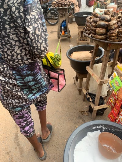 This media file shows a high-angle, eye-level shot of a market scene, likely Shagamu, Nigeria. The focus is on the lower body of a person wearing a vibrantly patterned wrap skirt and dark flip-flops, standing in a marketplace. In front of this person, a yellow bag filled with green produce is visible. Behind them, a pink and black bag rests on the ground.
The background reveals a bustling market environment. Several large dark basins are present, one filled with white powder and a brown scoop, and another empty. Wooden stalls hold dried fish, packaged spices, and other goods. A bicycle is also visible, leaning against a stall. The ground is paved with concrete. The lighting suggests daytime, possibly mid-morning or afternoon, with no indication of specific weather conditions. The scene captures the everyday activity of a marketplace, with goods for sale and people going about their business. No discernible emotions are conveyed, as the faces of individuals are not the primary focus. Some text is visible on the spice packets, including "SPICY" and "HOT PEPPER POWDER."