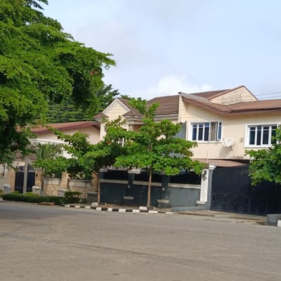 This is a photograph of a residential street in Gwarinpa, Nigeria, featuring a modern two-story house with beige-colored walls and a brown tiled roof. The house has multiple windows with white frames, and a satellite dish is mounted on the exterior wall. In front of the house, there is a black metal gate and a low wall with decorative pillars. Lush green trees and foliage are visible throughout the scene, partially obscuring the house. The street in the foreground is paved with asphalt and has a black and white striped curb. The sky is partly cloudy. There are no people or animals visible in the image.
