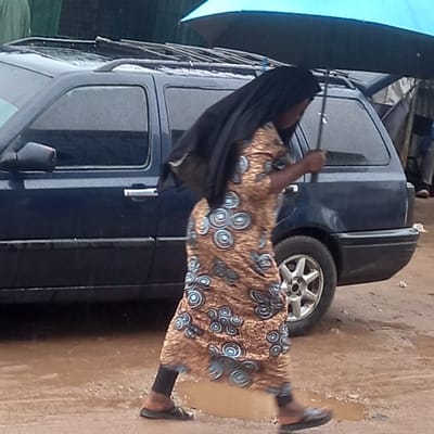 A woman is walking on a wet ground, holding a black umbrella beside a dark blue car on a street in Dadi Riba, Nigeria. The woman wears a brown and blue patterned dress over black leggings and black sandals. The car has a roof rack.