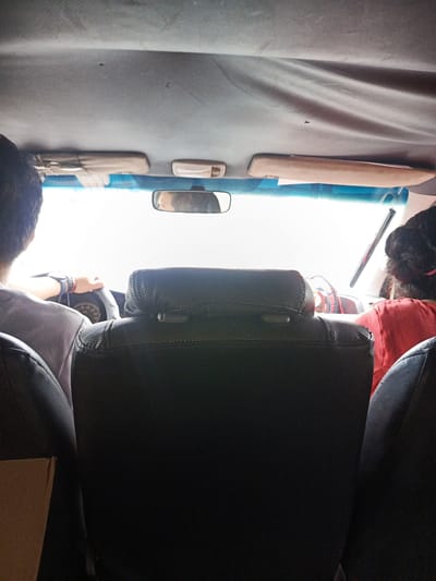 The image depicts the interior of a car, taken from the rear seats. The driver and a passenger are visible in the front seats. The driver is holding the steering wheel. A rearview mirror is present. The background, visible through the windshield, is bright and indistinct. The car's interior, including the seats and ceiling, appear to be worn. The location of the image is inferred to be the city of Quevedo, Ecuador.