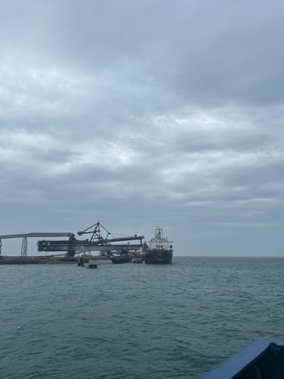 A wide shot captures a cloudy sky over a body of water, with an industrial port and a cargo ship visible on the horizon. The port features large loading cranes and structures. A cargo ship, partially docked, is on the right side of the frame. The water is choppy, reflecting the overcast sky. A blue structure, likely part of another vessel, occupies the lower right corner. This scene is located in the city of Uribia, Colombia.