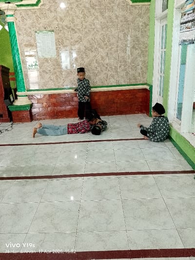 The image shows an interior room with tiled floors and walls. Three young boys are in the room. One boy is standing, facing the camera, with his hands at his side. Another boy is lying on his stomach with his head resting on a black object, facing the left side of the image. The third boy is sitting cross-legged on the floor, facing the camera. The room is a mosque located in Anjatan, Indonesia, at the coordinates provided. The room has a green wall on the right side with a white door and brown tiled walls on the left side. There are also white tiled floors with red lines running across the floor. The boys are all wearing similar black and white patterned clothing. The image was taken on April 17, 2023.
