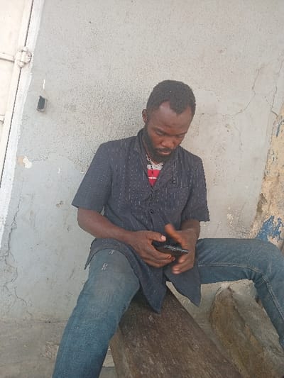 A man sits, dressed in jeans and a striped shirt, holding a mobile phone. He appears to be outside, leaning against a wall with peeling paint. His posture and the position of the phone suggest he's focused on the screen, possibly texting or browsing. The scene is simple, with the man as the central figure engaged in a digital activity within a basic, somewhat rundown setting.