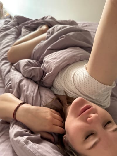 A fair-skinned individual is lying in a bed with their eyes closed. They are covered with a lavender duvet or comforter. The person is wearing a white shirt with small dots. One hand is near their head and has a dark-colored bracelet on the wrist. Part of their legs are visible underneath the comforter. The scene appears to be indoors, possibly in a bedroom with a wall visible in the background. There is no indication of this image being in Gornyi.