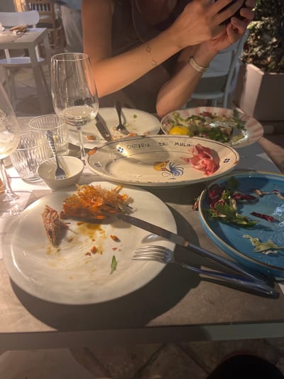 The scene captures a post-meal setting at a restaurant in Polignano a Mare, Italy. Four partially-eaten plates are visible, containing remnants of salad, cured meat, and seafood, specifically shrimp. Two sets of silverware rest on or near the plates. Multiple empty wine glasses are situated at the edge of the table. A person wearing bracelets is captured on the left, holding their hands to their face, possibly operating a phone. A partial view of another person is seen on the right. The setting appears to be an outdoor dining area.