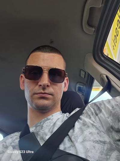 A frontal selfie captures a male individual, estimated to be between 25 and 40 years old, seated within the front passenger area of a vehicle. The man has closely cropped dark hair and visible light stubble on his face. He wears dark-tinted, gold-framed aviator-style sunglasses. His attire consists of a white t-shirt featuring a grey, mottled, abstract pattern. A black seatbelt spans diagonally across his chest and left shoulder. Beneath the seatbelt, a black strap, consistent with a crossbody bag, is visible over his left shoulder. The vehicle's interior headliner is grey fabric, and a beige plastic passenger grab handle is mounted above the window on the right side of the frame. Through the partially open window, an exterior yellow sign displays inverted characters "+1929!", indicative of fuel pricing at a gas station. A light blue sky is visible in the background through the window. The image contains a "Galaxy S23 Ultra" watermark in the lower-left corner. This scene is observed in Haskovo, Bulgaria.