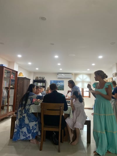 A group of people are gathered around a table indoors, likely in a residential or dining setting. A woman in a blue and white patterned dress is seated at the table, reaching forward with a utensil. Next to her, a man in a black suit jacket is seated, facing away from the camera. Several other individuals are visible around the table, some engaged in conversation or eating. In the foreground, a woman in a turquoise dress stands holding a plate and a fork, appearing to be eating. The room is well-lit with recessed ceiling lights and has several pieces of furniture, including a wooden cabinet filled with books and decorative items, and a large bookcase. Artwork hangs on the walls, and an air conditioning unit is mounted high on one wall. The flooring appears to be tiled. The overall atmosphere suggests a social gathering or meal. Based on the context of Ko Samui, Thailand, this scene could be taking place in a villa, a restaurant, or a private residence on the island.