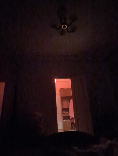 The image captures a dimly lit interior, possibly a room. In the upper center, a chandelier with a circular element is visible against a dark ceiling. The main focus is an open doorway in the middle distance, illuminated by a warm, orange light emanating from the room beyond. Through the doorway, a portion of a room is visible, featuring a wooden shelf unit with some items on it, and a person partially obscured by a pink garment. The foreground is very dark, obscuring most details. The location of Pkhvenisi, Georgia, is not indicated by any visual cues in the provided image.