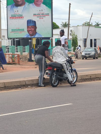 The image depicts a street scene in Dadi Riba, Nigeria, under an overcast sky. A large political billboard occupies the upper left, featuring three male figures. From left to right, the billboard displays "ASIWAJU BOLA AHMED TINUBU PRESIDENT FEDERAL REPUBLIC OF NIGERIA," "SEN MAL UBA SANI EXECUTIVE GOVERNOR KADUNA STATE 4+4," and "RT. HON. MUHAMMAD ABUBAKAR MAMADI KADUNA STATE CO-ORDINATOR RENEWED HOPE."
In the foreground on the right, a person wearing a white and black striped long-sleeve shirt with "RAPSTAR" text and matching striped pants is seated on a blue and black motorcycle. Next to the motorcycle, a person in a black short-sleeve top, grey sweatpants with white side stripes, and black sandals is standing, facing the seated individual. A third person, wearing a white graphic t-shirt and a black beanie, is partially visible behind the motorcycle driver, looking towards the left.
The road is asphalt with a single white dividing line. A concrete curb separates the road from a dirt and gravel shoulder, where some debris is visible. On the far left, a person in a dark blue garment is walking away from the camera. Orange construction barriers and several black water storage tanks are visible behind the walking individual.
In the mid-ground right, a silver Toyota Matrix with a visible "KADUNA" license plate is on the road, alongside a red motorcycle. A person in a light-colored traditional garment is standing near the red motorcycle. White, modern-style buildings with blue-tinted windows and external air conditioning units are in the background. Palm trees and utility poles are also present.