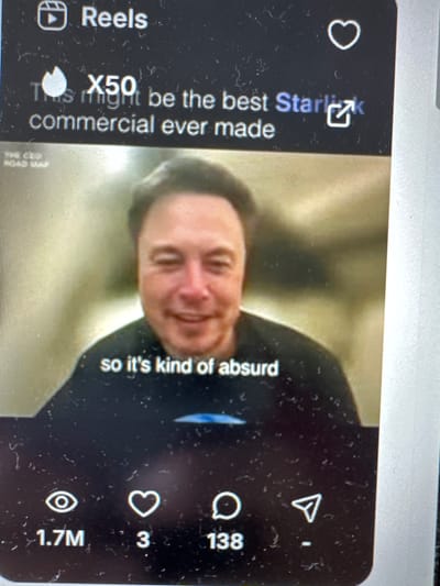 The image shows a screenshot of a social media post, likely from a platform like Instagram Reels or TikTok. The post features a video of Elon Musk speaking, with text overlays. The top of the screen has "Reels" and a play button icon, suggesting it's a video format. Below this, there's text that reads "X50" and "This might be the best Starlink commercial ever made". In the video itself, Elon Musk is shown with a microphone and wearing a black shirt. The subtitle below him says, "so it's kind of absurd". The bottom of the screen displays engagement metrics: "1.7M" views (indicated by an eye icon), "3" likes (indicated by a heart icon), and "138" comments (indicated by a speech bubble icon), and a send icon. The image is a photograph of a screen, with some dust or scratches visible on the surface. There is no indication of Barnaul, Russia, or any specific city location within the image.