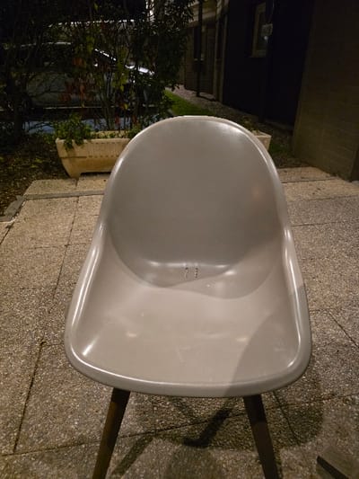 A light-gray chair, seen from an overhead angle, dominates the right side of the frame, its smooth, curved surface contrasting with the textured, stone-like ground beneath. To the left, a dark building facade recedes into the background, partially obscured by foliage. The lower-left portion of the image features a cluster of trees and greenery, softened by the dim light. The lighting casts shadows across the ground, highlighting the chair's form and the surrounding environment.