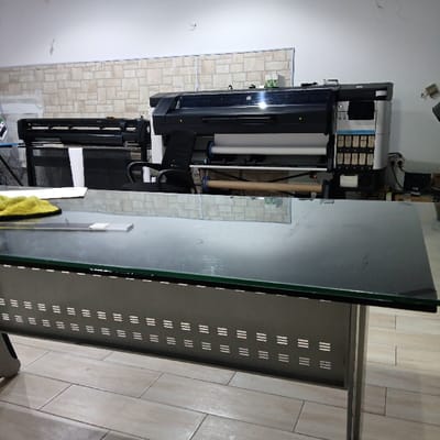 A brightly lit room features a modern glass-topped table, reflecting the scene behind it. Behind the table, two large format printers dominate the background, positioned against a brick-patterned wall. One printer is black, and the other is a multi-toned device. A black office chair is partially visible, and various equipment, including power supplies, suggest a printing or design workspace. The floor is tiled in neutral tones.