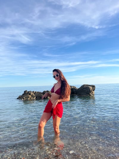 A woman stands in shallow, clear water near a rocky outcrop under a blue sky with scattered white clouds. She is wearing a burgundy bikini and a red sarong, and white sunglasses. Her long, dark hair is down. The sea is calm, and the seabed is visible with pebbles and stones. The scene appears to be a beach or coastline.