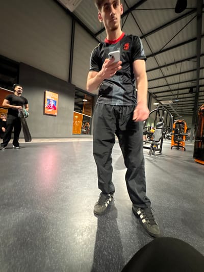 A man in athletic attire stands indoors, holding a phone. He wears black pants and Adidas sneakers. Gym equipment is visible behind him, including weight machines and an orange Matrix trainer. In the background, a poster with a tennis player and the word "HEAD" adorns the wall, and two other men are seen carrying equipment. The setting appears to be a gym or training facility. The photo is taken at an angle, emphasizing the industrial ceiling structure.
