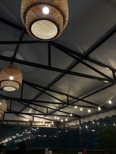 The image depicts the interior of an outdoor dining establishment, likely a cafe or restaurant, with a covered ceiling supported by a dark metal frame. Two large, woven bamboo pendant lamps hang from the ceiling, casting a warm glow. String lights with exposed bulbs are also visible along the perimeter of the covered area. A transparent plastic curtain hangs along the sides, offering some enclosure. In the background, a person is seated at a table, facing away from the camera, and a potted plant is visible on the right. The overall ambiance suggests a cozy, dimly lit evening setting. The location of this establishment is in the city of Neftekamsk, Russia.