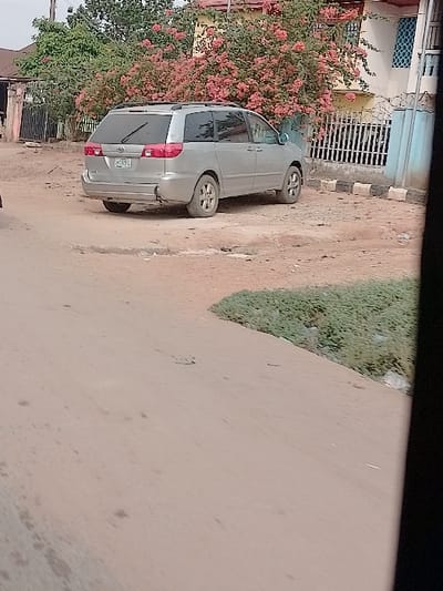The scene depicts a static outdoor environment in Orie Uratta, Nigeria, under clear daytime lighting. A silver Toyota Sienna minivan, with visible license plate characters "BAY 782", is parked on unpaved light brown earth, angled towards the right. The vehicle's rear bumper shows damage, with a missing section on the lower left. Behind the minivan, a prominent bush with numerous pink flowers (consistent with bougainvillea) partially covers a light yellow building. A white metal fence, topped with barbed wire, is situated in front of the building. To the immediate right of the minivan, square concrete blocks line what appears to be a property boundary. Further right, a light blue building with a patterned blue window is visible. The foreground consists of an unpaved dirt path, with a patch of green ground cover visible on the right. No individuals or active events are depicted.