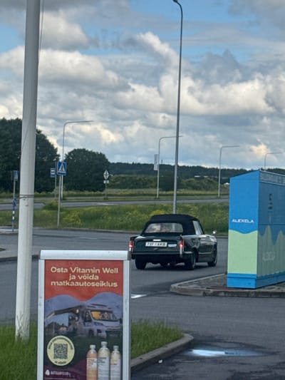 In Tiskre, Estonia, a black convertible car with license plate "737 AKB" is parked near a bright blue and green kiosk labeled "ALEXELA." A large red advertisement board stands in the foreground, featuring text in Estonian about Vitamin Well and a camper van. The scene is set outdoors with a cloudy sky overhead and greenery in the background, suggesting a roadside or parking area. No people are clearly visible in the image.