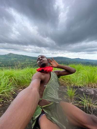 This is a vertical, eye-level, first-person perspective photograph taken outdoors in Achara, Nigeria. The scene features a young Black man lying on his back on a rocky outcrop, surrounded by lush green grass and vegetation. The sky is overcast and filled with dramatic, heavy grey clouds, suggesting a stormy or impending weather event. In the background, rolling hills covered in green foliage descend into a valley, with some small structures visible in the distance, hinting at a rural or semi-rural landscape.

The man is wearing a red band around his neck and is partially draped in large green leaves, possibly as a makeshift garment or costume. His face is turned towards the camera, and he appears to be smiling or smirking, with white markings or paint on his forehead and cheeks. One of his arms is raised, with his hand open, in a gesture that could be a wave or a pose. The lighting is dim due to the cloudy sky. The overall mood is somewhat dramatic and naturalistic, with the imposing clouds contrasting with the vibrant green of the landscape and the man's relaxed posture. No visible text is present in the image.
