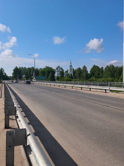 The image shows a road on a bridge with a metal guardrail on the left side. On the right side of the road, there is a decorative metal fence, a green building, and a white church with a spire in the background. The road is paved and there are cars on it. The sky is blue with white clouds. Trees line the background. The scene appears to be in the city of Kady, Russia, as indicated by the architecture of the church.