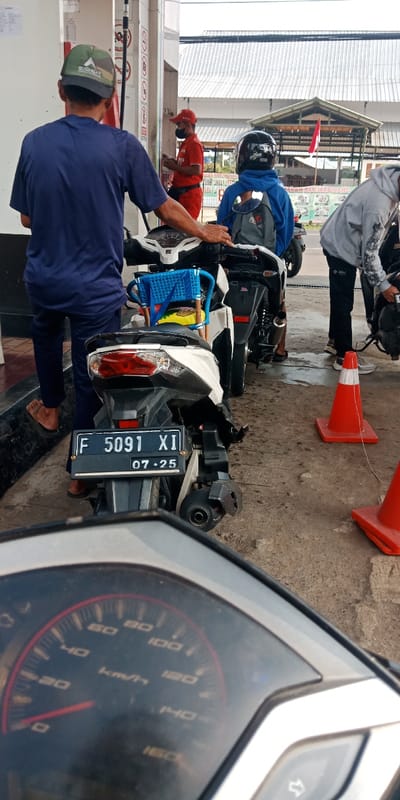 The image depicts a daytime scene at a Pertamina gas station in Cijedil, Indonesia. The perspective is from the front of a stationary motorcycle, with its speedometer showing a maximum of 160 km/h visible in the foreground. Directly ahead, the rear of a white Honda Vario scooter is visible, displaying license plate F 5091 XI with an expiration date of 07-25. A blue plastic child seat is attached to the white scooter's floorboard.
A man wearing a dark blue shirt, dark pants, and a green camouflage cap with an "EIGER" logo stands beside the white scooter, facing a fuel pump. His right hand rests on the scooter's handlebar. Behind him, another individual in a blue hoodie and black helmet (with "NHK" branding) sits on a black motorcycle, equipped with a grey backpack. A gas station attendant, dressed in a red uniform, red cap, and black face mask, stands near a fuel pump, looking at a mobile phone. To the right, another person in a light grey hoodie is bending over a third motorcycle. Two orange traffic cones are placed on the concrete ground.
In the background, a white canopy covers the fuel pumps. Beyond the gas station structure, buildings with corrugated metal roofs and partial signage, including the words "MAKAN BANJARDINANG," are visible. An Indonesian flag, red and white, flies on a pole. Overhead utility lines are present.