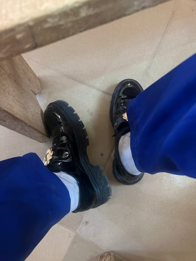 The image presents a close-up, downward-angled view of the lower legs and feet of an individual in an indoor setting. The individual is wearing royal blue trousers and white ankle socks. On their feet are a pair of black, patent leather-style loafers with thick, lugged soles. Each shoe features a prominent gold-colored metal buckle or ornamentation across the instep. The individual's left foot is positioned closer to a light brown wooden structure, likely the leg of a table or desk, which occupies the upper left quadrant of the frame. Both feet rest on a floor composed of light beige square tiles with visible grout lines. Reflections are present on the glossy surface of the shoes, indicating ambient indoor lighting. Specific visual identifiers to ascertain a precise location within Birnin Kebbi, Nigeria, are not discernible from the provided image. The individual appears to be seated.