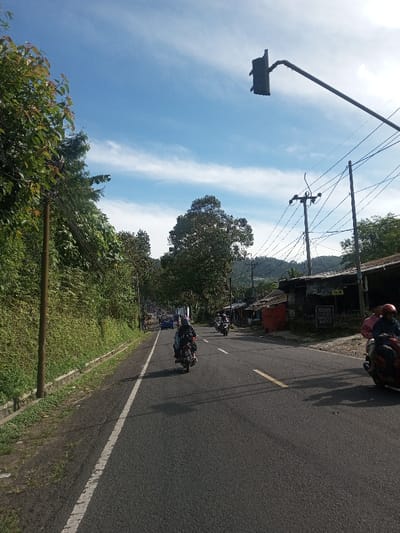 The image depicts a daytime scene on a paved road in Phonengam, Laos, under a clear blue sky with scattered white clouds. The asphalt road features a solid white line marking the left edge and a dashed yellow line dividing lanes.

On the left side of the road, a steep, vegetated embankment with dense green foliage extends upwards. A wooden utility pole with multiple power lines is visible along this embankment.

On the right side of the road, a series of rudimentary roadside structures or stalls are present, constructed from varied materials including corrugated metal and tarpaulin. Signs are visible on these structures, one displaying text. Two metal utility poles support numerous intertwined electrical wires overhead. A black, rectangular traffic light is mounted on a metal pole extending over the road from the right.

Multiple motorcycles are traveling on the road. In the foreground, a dark motorcycle with a rider wearing a dark helmet and jacket, and a passenger, moves away from the viewer. Further down the road on the right, another motorcycle with a single rider is visible. On the far right, a red motorcycle with a rider in a red helmet and pink shirt, accompanied by another standing individual, is partially captured. Additional motorcycles and a blue car are visible in the distant traffic.

The background shows lush green trees and rolling hills or mountains under the bright sky. Shadows are cast across the road from the right, indicating sunlight from the left.