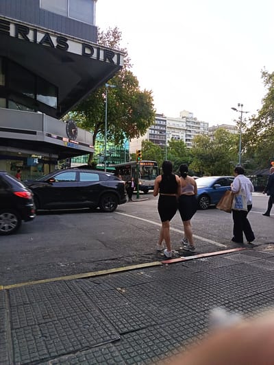 The image shows a street scene in Montevideo, Uruguay, identified by the "FERIAS DIRI" sign on a building in the background. The street intersection has cars, a bus ("110 MANGA") , and pedestrians. Two women in athletic wear are standing on the crosswalk, and another woman carrying a bag is walking next to them. A man in a suit is walking on the far right. A black car and a blue car are driving through the intersection. The day is overcast.