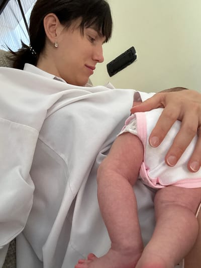 A woman with dark hair is seen lying down in a white shirt, holding a baby. The baby is wearing a white outfit with pink trim. A remote control is attached to the wall in the background. The scene appears to be indoors. The location is unknown.