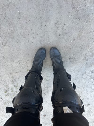 The image shows a first-person perspective of two legs wearing black motorcycle boots standing on a concrete surface. The boots appear to be slightly worn and dusty. The concrete surface is light grey with small pebbles and imperfections visible. The location is not identifiable as Cherliany, Ukraine from the image alone.