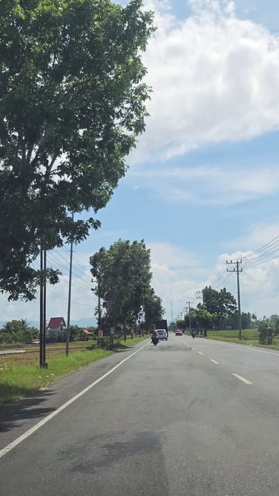 The image depicts a road scene in Kampungjumpa, Indonesia, showcasing a two-lane asphalt road with a white line separating the lanes. Trees line the left side of the road. Power lines stretch across the sky. A vehicle and a motorcycle are visible on the road. The sky is blue with scattered clouds. Grass and a house are visible on the roadside.