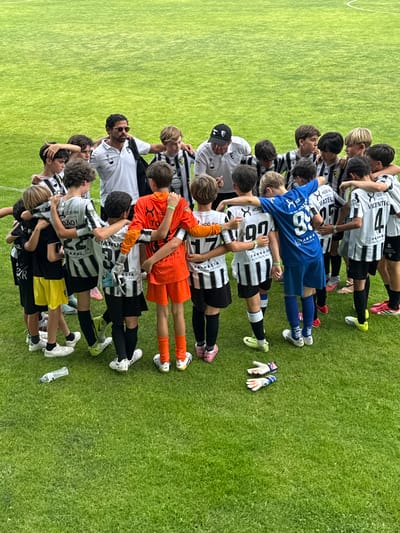 An outdoor, high-angle, full-shot photograph captures a youth soccer team, including two adult coaches, in a huddle on a vibrant green grass field in Santa Maria da Feira, Portugal. The team, predominantly wearing black and white striped soccer uniforms, stands in a circle, arms around each other's shoulders. Two adults, likely coaches, are positioned on the left and right sides of the circle, facing inwards towards the players. One coach, on the left, wears a white polo shirt and sunglasses, with his arm around a player. The other coach, on the right, is partially visible, wearing a white shirt and a black baseball cap. Scattered on the grass are a water bottle and a pair of goalkeeper gloves. The backdrop consists of the meticulously maintained green soccer field, marked with white lines of a pitch.