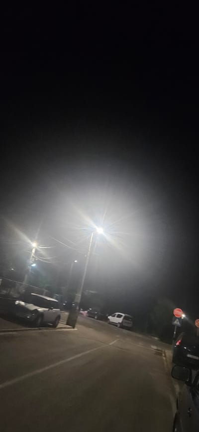 The image depicts a nighttime scene on a street, likely in or near Kostenets, Bulgaria, due to the presence of visible Bulgarian stop signs. The primary light source appears to be a bright street lamp in the center of the frame, creating a significant lens flare and illuminating the misty or foggy atmosphere. Several parked vehicles are visible: a white SUV-like vehicle on the right side of the road, a darker colored car partially visible on the left, and other vehicles further down the street. The road surface is paved, with visible lane markings. Power lines and poles are also present, typical of a street scene. The overall impression is one of low visibility due to the darkness and atmospheric conditions.