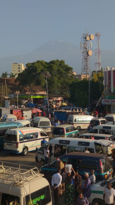 A bustling street scene showcases rows of white minibuses packed with people. Some individuals stand near the vehicles, possibly interacting or boarding. In the background, a lush canopy of green trees fills the space, with cell phone towers standing tall on the left and a snow capped mountain behind them. Buildings line the street, blending into the vibrant backdrop of this busy urban area.