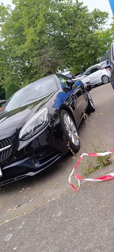 The image shows a black Mercedes-Benz convertible parked on an asphalt surface, likely a street or parking lot, with a wooded area and other vehicles in the background. The car is positioned at an angle, with its front left quarter prominently displayed. Red and white caution tape is visible on the ground in the foreground, suggesting a cordoned-off area. The environment appears to be outdoors, with trees and foliage indicating a possibly suburban or park-like setting. The scene is devoid of people, and the overall impression is one of a static, parked vehicle. There is no specific indication of the city of Waiblingen, Germany, within the visual elements of the image itself.