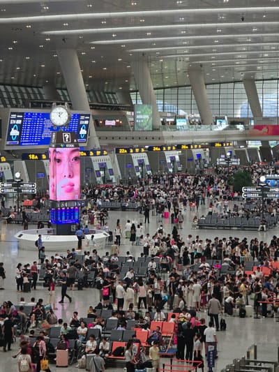 The image depicts a crowded interior of a large, modern transportation hub, likely a train station or airport, in Shangcheng District, China. The space is vast and bright, with a high, arched ceiling featuring numerous light fixtures and large, cylindrical support columns. Digital display boards are visible throughout, showing departure and arrival information, as well as advertisements.
A prominent central feature is a large, vertical LED screen displaying a close-up of a person's face wearing glasses. Below this screen, a digital information kiosk with a question mark symbol is positioned. Adjacent to the large screen, a traditional analog clock with a white face and Roman numerals is mounted on a digital board.
Hundreds of people are present, many seated on rows of blue and orange chairs arranged in waiting areas. Others are standing, walking, and interacting within the space. Most individuals are dressed in casual attire. Some are pulling luggage, indicating travel. Several individuals are looking at their phones, while others are engaged in conversation or observing the information boards.
The floor is a polished, reflective surface. The environment is clean and well-lit, with a mix of artificial and natural light filtering in from large windows in the background. The overall impression is one of a busy, organized, and functional public space.