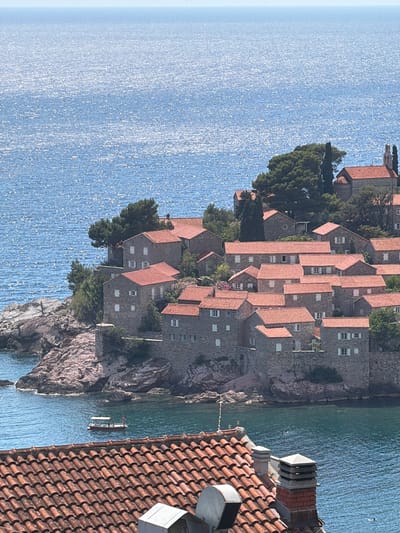 The image shows a picturesque view of the island town of Sveti Stefan, Montenegro, with the Adriatic Sea in the background. The island is densely packed with stone houses topped with red tile roofs, nestled amongst lush greenery. The sun glints off the deep blue water, creating a shimmering effect. In the foreground, the terracotta tiled roof of another building is visible, with a chimney stack. A small boat with two people on board is visible in the turquoise water near the island. The scene is bathed in bright sunlight, suggesting a clear, warm day.