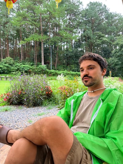 A man sits outdoors in what appears to be a park or garden area in Ban Ang Ka Noi, Thailand. He is wearing a tan t-shirt, olive-colored shorts, a bright green hooded rain jacket, and brown slides. He has dark curly hair, a mustache, and a beard. He is wearing a white necklace. He is seated on a wooden surface with his legs crossed. The background features a dense forest of tall pine trees, lush green vegetation, and flowering bushes with purple and red flowers. There are also some white flowers visible. The ground is covered in small grey gravel. Two red and yellow lanterns are visible in the upper left corner of the image, suggesting a festive or decorated area. The weather appears to be overcast, possibly after a rain shower, as indicated by the presence of water droplets on the rain jacket.