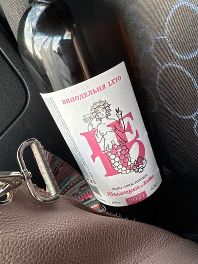 The image shows a close-up of a wine bottle with a pink and white label. The label has text in Russian and a graphic of a woman holding grapes. The bottle is dark glass and appears to be a rosé wine. The bottle is leaning against a textured surface, possibly a car seat, and a brown leather purse is in the foreground. The scene does not provide any information or context that would allow for the determination of its location in the city of Veliky Novgorod, Russia.