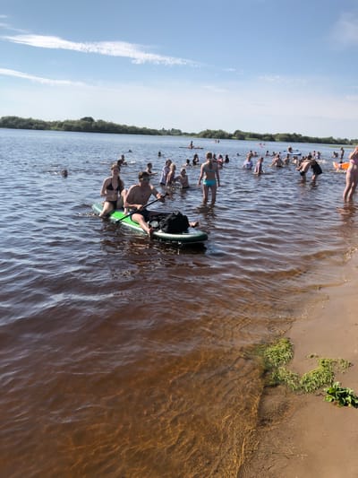 This media file captures a lively scene of people enjoying a sunny day at the waterfront. The image features a gathering of individuals, many of whom are in swimwear, engaged in various water activities. A prominent group is seen on paddleboards, with some individuals actively paddling while others are seated. Several people are wading or standing in the shallow water near the shore, while others are further out in the lake. The water appears to be calm with gentle ripples reflecting the sunlight.

The setting is a natural waterfront, with a sandy or muddy shore visible on the right side of the image. Green vegetation, possibly reeds or grass, grows along the edge of the water. The background shows a dense line of trees, suggesting a forested area. The sky is a clear, bright blue with a few wispy clouds, indicating fair weather. The sunlight suggests it is daytime, likely afternoon, given the brightness and the presence of shadows. The atmosphere appears relaxed and joyful, with people socializing and engaging in recreational pursuits.

There are no visible text elements in the image. The scene conveys a sense of summer recreation and community enjoyment of an outdoor water environment.
