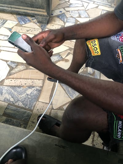 A person is sitting on a step, holding a smartphone in their hands. The phone is green and is being used with the right hand. The person is wearing dark shorts with patches, one of which reads "DSQ2". A white charging cable is connected to the phone and trails off to the left. The person is also wearing black sandals. The ground is tiled with a mosaic pattern of various colored stones. The background is dark and out of focus. The scene appears to be outdoors, possibly in a public area. There is no specific indication of the city of New Stadium, Nigeria, in the image.