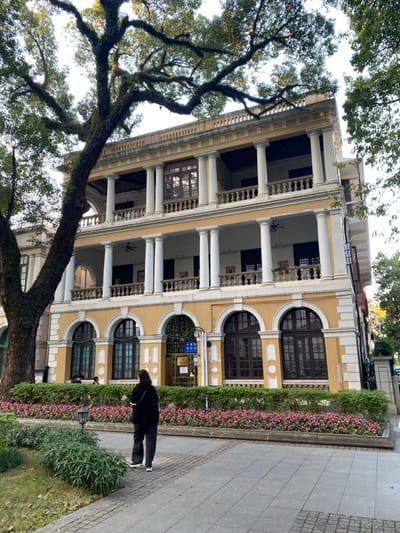 A large, multi-story building with a distinctive architectural style dominates the scene, partially obscured by a mature tree in the foreground. The building features a light yellow facade adorned with prominent white columns supporting multiple levels of open balconies or verandas, some with white balustrades. The ground floor displays large, dark-framed arched windows, while the upper levels have rectangular windows and visible ceiling fans within the covered balcony spaces.

On the left, a large, old tree with a thick, gnarled trunk and extensive branches covered in moss or lichen fills a significant portion of the frame, its green foliage silhouetted against a bright, clear sky.

In front of the building, a well-maintained garden bed featuring green shrubs and small pink flowers lines a paved sidewalk. A person, dressed in dark clothing and possibly carrying a backpack, stands on this sidewalk, facing away from the camera and looking towards the building.

The image is well-lit, indicating daytime. A blue sign visible on the building's entrance clearly displays Chinese characters "出入口" (chū rù kǒu), which translates to "entrance/exit." The setting suggests an urban environment, consistent with the specified location of Guang Zhou Shi, China, showcasing a blend of natural elements and historical architecture.