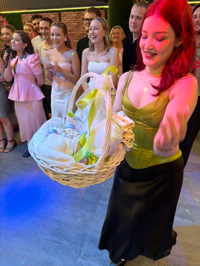 In Chernushka, Russia, a group of young adults are gathered indoors, likely at a celebration or party. The foreground is dominated by a young woman with vibrant red hair, illuminated by pink and red lighting, wearing a lime green corset-style top and a long black skirt. She is smiling and holding a wicker basket filled with gifts, including what appear to be baby items, wrapped in white fabric and adorned with yellow and green ribbons. Behind her, several other individuals are visible, including a woman in a pink outfit, a woman in a strapless white top, and various men and women in casual attire. The background features a brick wall and a dense green hedge. The floor is tiled and reflects the blue and green lighting.