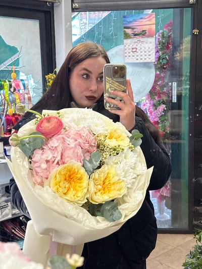 A brunette young woman poses with a bouquet of flowers in front of a window or mirrored surface, taking a selfie with her smartphone. She is wearing a black jacket. The bouquet features a variety of pastel-colored flowers including pink hydrangeas, yellow roses, white chrysanthemums, and a single red ranunculus. The bouquet is wrapped in a light-colored paper and tied with a ribbon. The reflection shows a calendar dated March 2022 and other objects suggesting a floral shop or similar setting. Part of an OCR text suggests "ALAND CAMARGO 720". The location of the city of Liepaja, Latvia is not evident in this image.