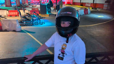 The image shows a child wearing a black helmet and a white t-shirt with Bart Simpson printed on it. The child is standing on a go-kart track. The track has yellow and blue markings and is lined with red and white barriers. In the background, there are several go-karts and what appear to be Coca-Cola advertisements. The overall scene suggests an indoor go-karting facility, possibly at an amusement park or entertainment center. There is no specific indication within the image that places the location in Östersund, Sweden; this information is external to the visual content.