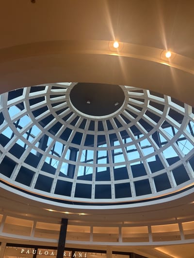 The image shows the interior of a shopping mall, likely in Wrocław, Poland, with a focus on the domed ceiling. The ceiling is a geodesic dome constructed with white structural elements and dark panels, allowing glimpses of the blue sky through its sections. Two circular lights are visible on an overhead curved structure. Below the dome, a circular balcony or walkway is visible, with several illuminated rectangular openings. At the bottom of the image, the signage "PAULO A RIANI" is partially visible, suggesting a retail store. A dark vertical pillar is also present in the lower part of the frame. The scene is brightly lit, with the warm tones of the interior lighting contrasting with the cooler tones of the sky.