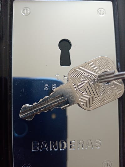 A close-up, high-angle photograph presents a shiny, metallic lock plate with a key inserted into its keyhole. The highly polished surface of the lock plate reflects a distorted, dark blue, textured area, likely the object used to capture the image. The key, made of a bright silver-colored metal, prominently displays an intricate, repetitive geometric pattern on its bow, consisting of chevron and zig-zag lines. A central circular design on the key's handle contains a stylized logo, possibly an abstract "TP" or "M". The bitting of the key is clearly visible as it enters the black keyhole.

Engraved text on the lock plate includes a partially obscured phrase above the keyhole, which appears to be "THE SECURITY," with "S E C" being the most discernible letters. Below the key and the keyhole, the word "BANDERAS" is clearly etched into the metal. Small, circular, raised markings resembling stylized double 'D's are visible in the top corners of the lock plate, possibly decorative elements or screw covers. The image captures the functional and aesthetic details of a locking mechanism, set in the context of Elektrostal, Russia.