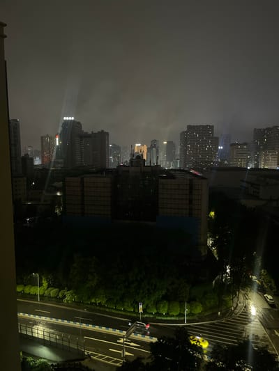 This detailed description pertains to an image captured at night within Jiulongpo District, China, depicting an urban landscape under a heavily overcast or foggy sky.

The scene presents a high-angle view of a city, dominated by numerous multi-story buildings, some illuminated from within or by exterior lighting. The sky is a dark, uniform gray, indicating a low cloud ceiling or significant atmospheric haze. Several distinct vertical light beams emanate upwards from specific high-rise structures, scattering through the atmospheric conditions.

In the mid-ground, a cluster of prominent buildings includes a tall structure with a vertical illuminated sign displaying Chinese characters, and an adjacent building exhibiting what appears to be a red-glowing horizontal sign. Near the center, large, box-like structures, possibly under construction or renovation, are covered with scaffold-like sheathing. Further in the background, a dense urban skyline is visible, characterized by numerous illuminated windows across various high-rise buildings, including one with a distinct golden-orange illuminated top.

The foreground features a complex road network. To the lower left, a multi-lane elevated road or bridge is present, distinguished by white and yellow lane markings and a blue-and-white barrier system along its edge. The road surface appears wet, reflecting ambient light. To the lower right, a curving street-level intersection or road segment is also wet and reflective. A bright yellow vehicle, exhibiting motion blur, is actively traversing this curving road segment, while a lighter-colored vehicle is stationary or moving slowly further along the street.

Extensive dark green foliage and trees are visible, particularly in the lower half of the image, lining the roads and filling interstitial spaces between urban structures. Several streetlights are active, casting localized illumination on sections of the greenery and road surfaces. No explicit human figures are discernible in the image.